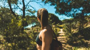 A woman with a ponytail wearing a dark sports bra and backpack stands on a sunlit forest trail, looking into the distance, surrounded by trees and natural greenery under a bright blue sky.