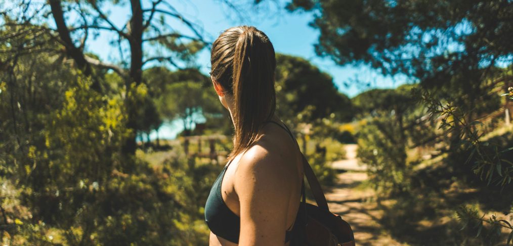A woman with a ponytail wearing a dark sports bra and backpack stands on a sunlit forest trail, looking into the distance, surrounded by trees and natural greenery under a bright blue sky.