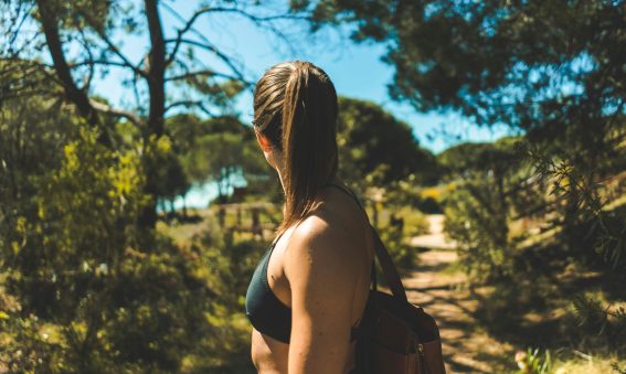A woman with a ponytail wearing a dark sports bra and backpack stands on a sunlit forest trail, looking into the distance, surrounded by trees and natural greenery under a bright blue sky.